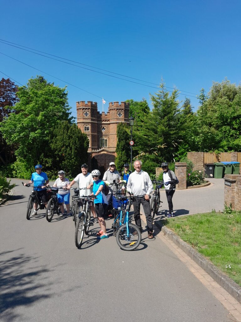 Standing cyclists beside their bikes, posing for a photo in front of double red-brick towers amongst trees