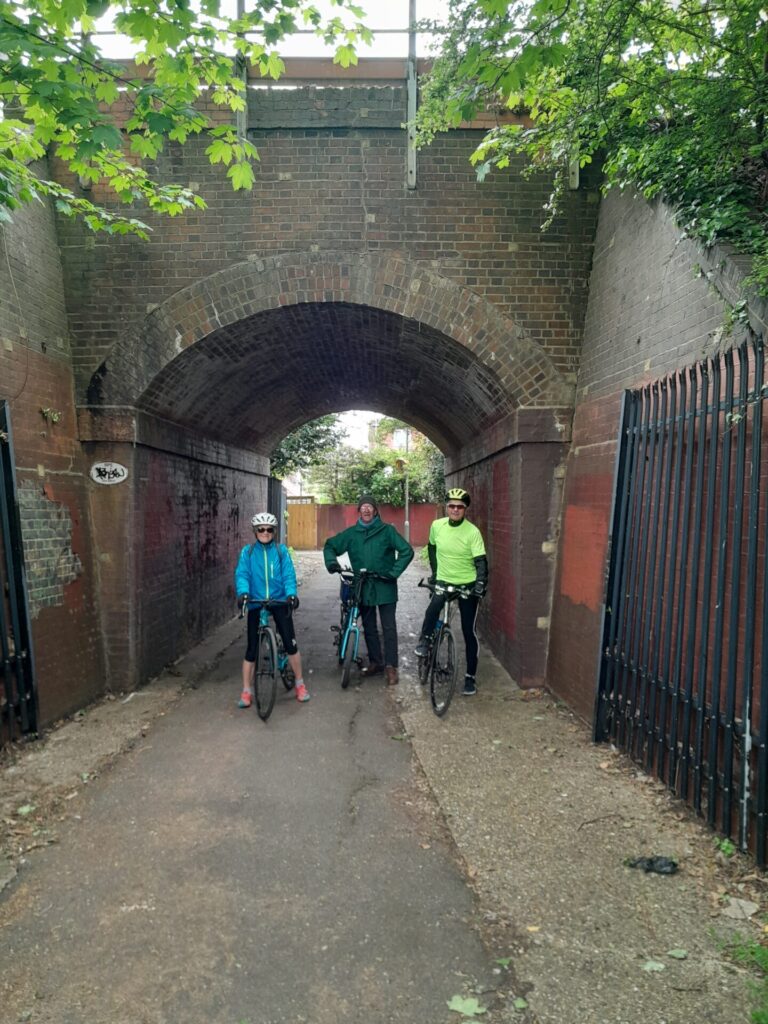 Standing cyclists with their bikes on a path, posing for a photo beneath a brick railway bridge arch