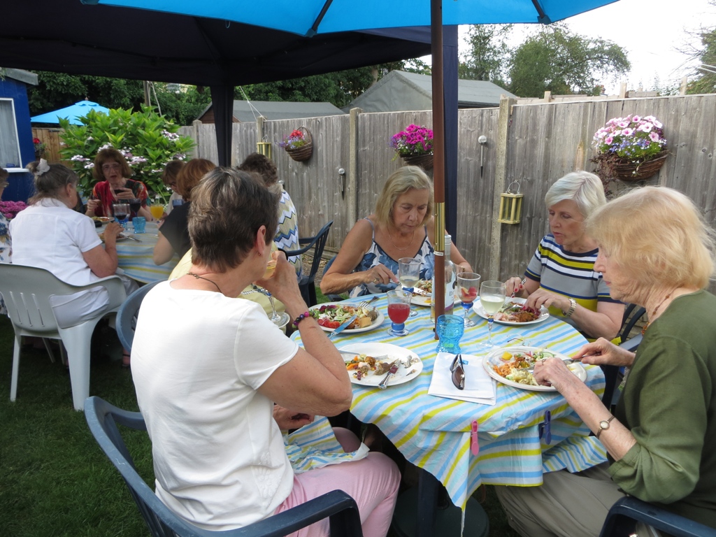 People seated around tables under sunshades in a back garden, eating and drinking